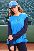 A woman wearing a blue and navy athletic long-sleeve shirt and matching blue cap stands on a baseball field with stadium seats in the background. The shirt features a bright blue body with navy sleeves and a green-and-blue chest logo.
|M1