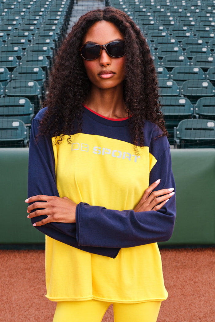 A woman with curly hair and sunglasses stands on a baseball field wearing a two-tone athletic long-sleeve shirt and matching yellow pants. The shirt is bright yellow on the front with navy sleeves and collar, featuring 'DB SPORT' printed across the chest.
|M1