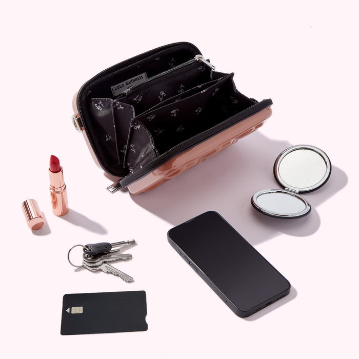 A rose gold Lulu Guinness purse is open, revealing its interior compartments. Beside it are a lipstick, a compact mirror, a smartphone, and a set of keys and a credit card. All items are on a white background.
