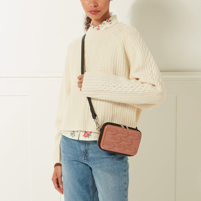 Woman wearing a cream-colored cable knit sweater and blue jeans, carrying a small blush-pink crossbody bag with a lip design.