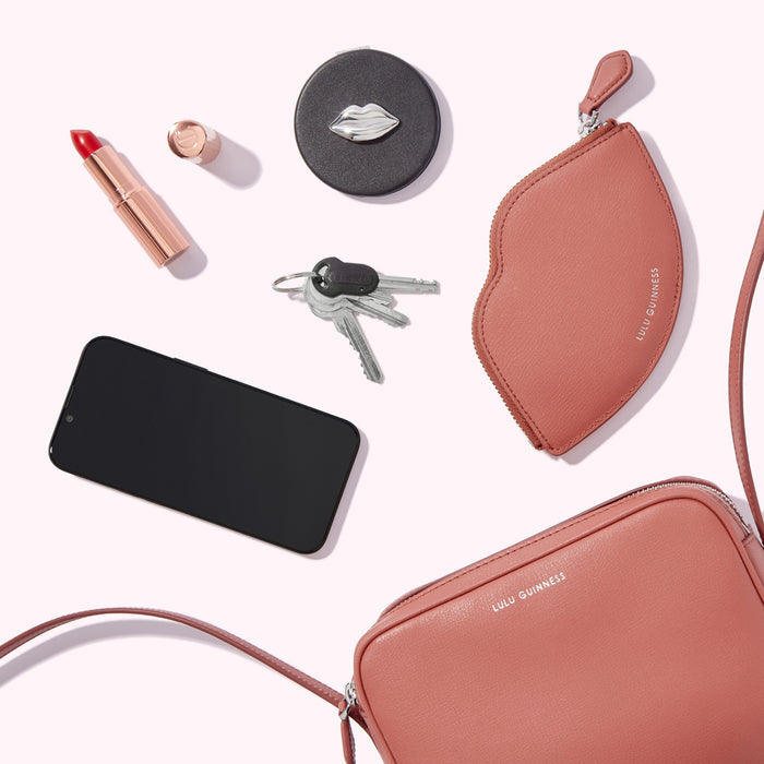 A flat lay of a Lulu Guinness crossbody bag, lip-shaped coin purse, compact mirror, lipstick, keys, and smartphone on a white background.