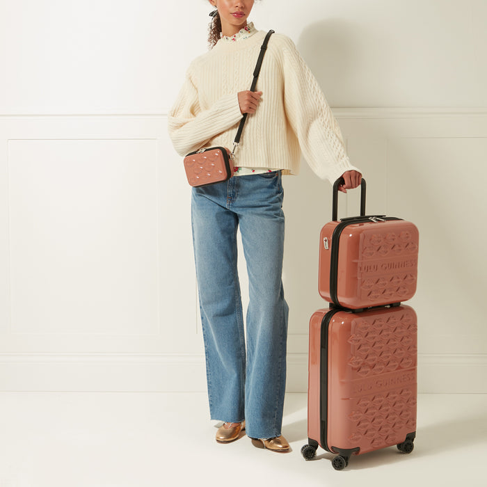 Woman in cream sweater and jeans holding a dusty rose Lulu Guinness suitcase set and crossbody bag against a white background.