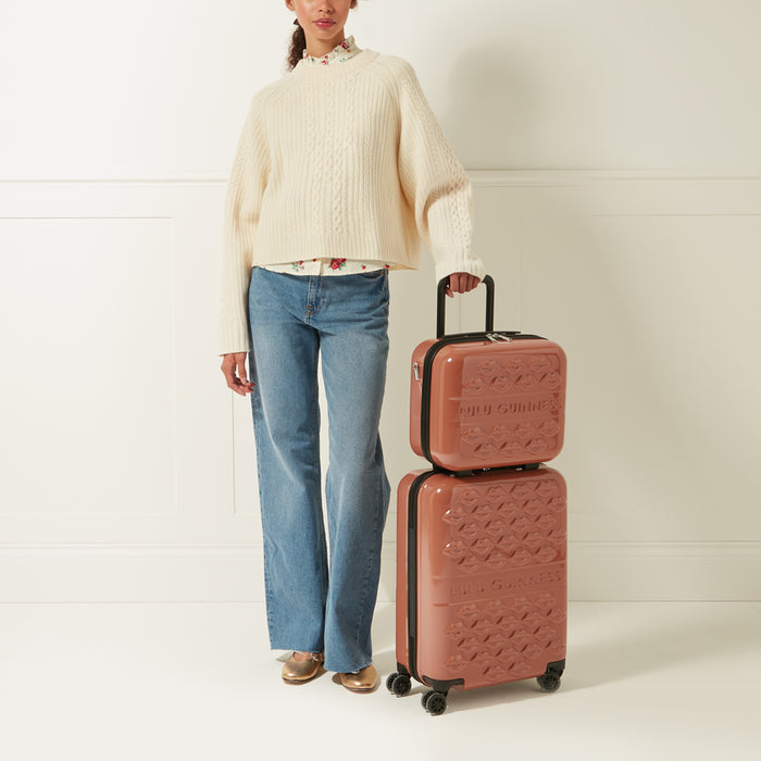 A woman in light cream sweater and blue jeans stands next to a dusty rose Lulu Guinness two-piece luggage set against a white background.