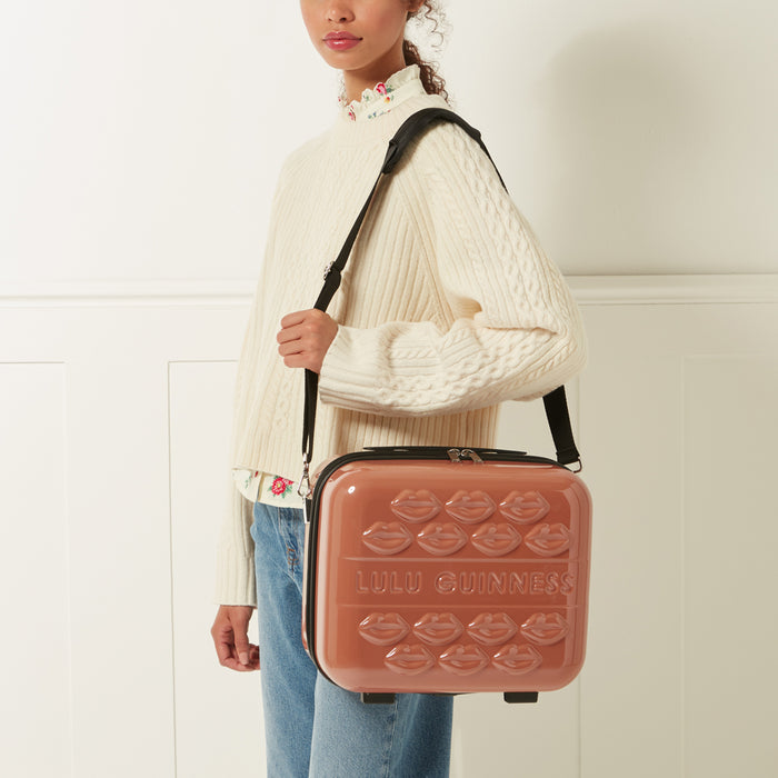 A woman carries a dusty rose Lulu Guinness makeup case with a black shoulder strap. The case is decorated with embossed lips.