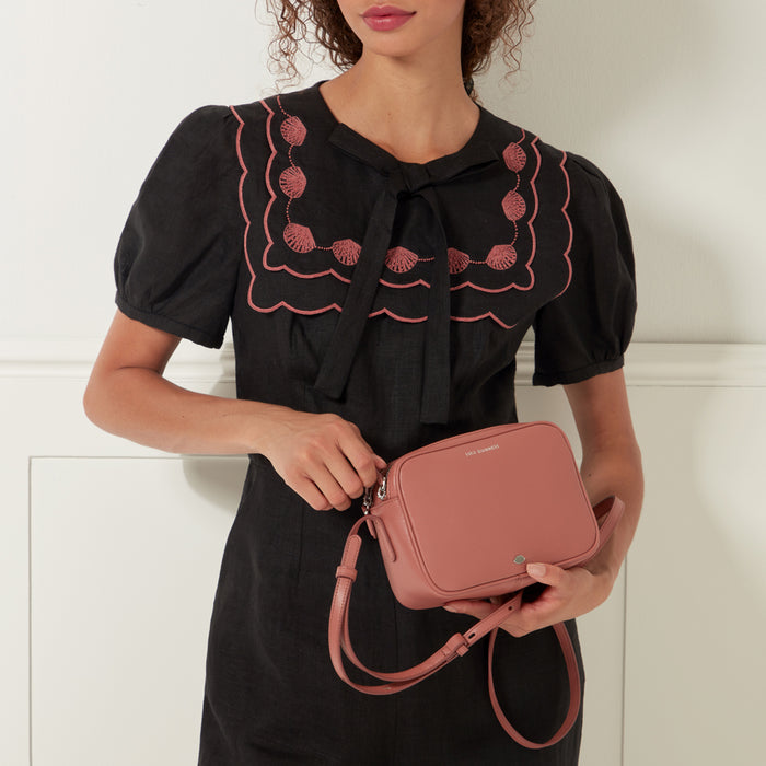 A woman in a black dress with pink shell embroidery holds a dusty rose-coloured Lulu Guinness crossbody bag.