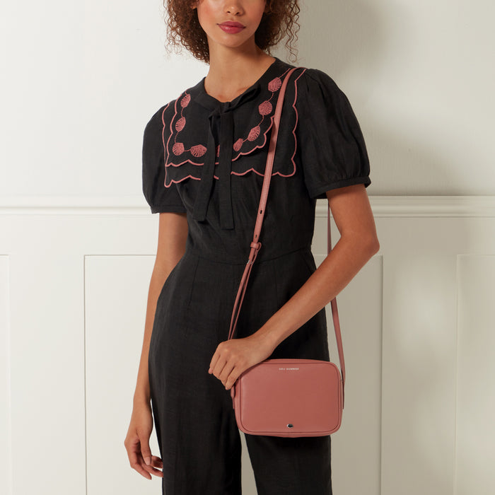 A woman in a black dress with pink embroidered collar holds a dusty rose-coloured crossbody bag. The background is off-white.