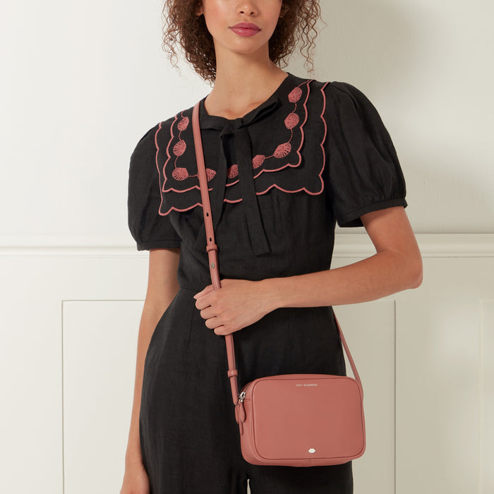 A woman in a black dress with pink embroidery holds a dusty rose-coloured crossbody bag.