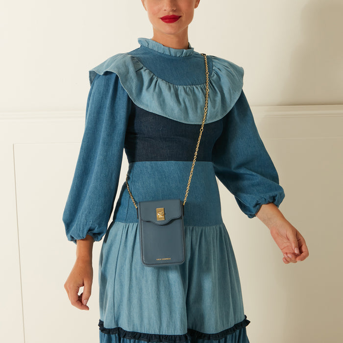 A woman in a blue denim dress models a dark-blue leather crossbody phone bag with gold accents.