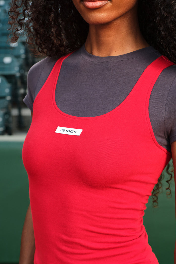 A close-up of a woman wearing a bright red athletic tank top over a dark gray short-sleeve shirt. The red tank top has a scoop neckline and a small white rectangular chest label that reads 'DB SPORT'.
|M1