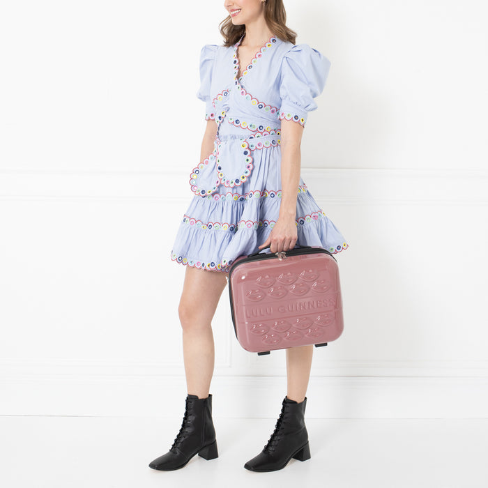 Woman in a light blue dress holding a pink Lulu Guinness lipstick-print suitcase against a white background.