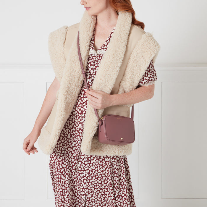 Woman in a burgundy floral dress and cream shearling vest holding a dusty rose crossbody bag.