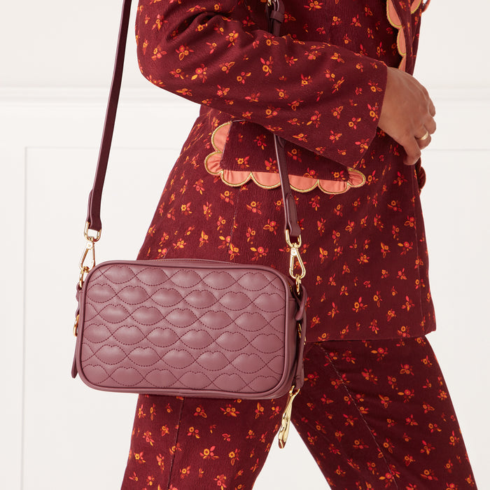 A mauve quilted crossbody bag with gold hardware hangs from the shoulder of a woman wearing a burgundy floral pantsuit.