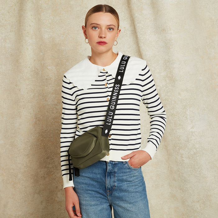 Woman wearing a striped shirt with a green bag with 'Lulu Guinness' on the strap, standing against a beige background