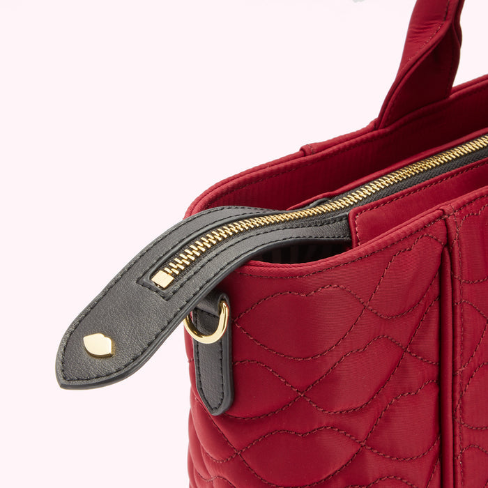 Close-up of a red quilted handbag's zipper and top handle. The zipper pull is black leather with gold hardware.