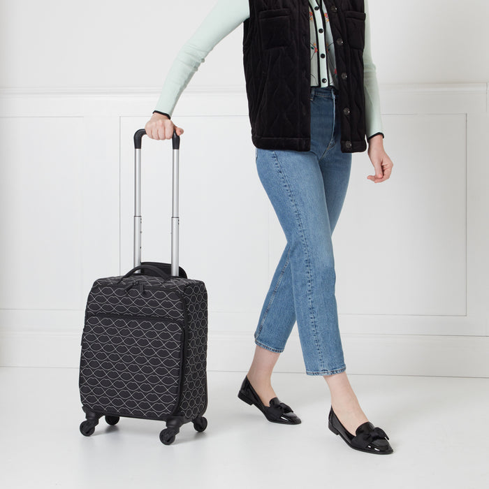 A woman in blue jeans and a black vest pulls a small black suitcase with a white lip print design on a white background.