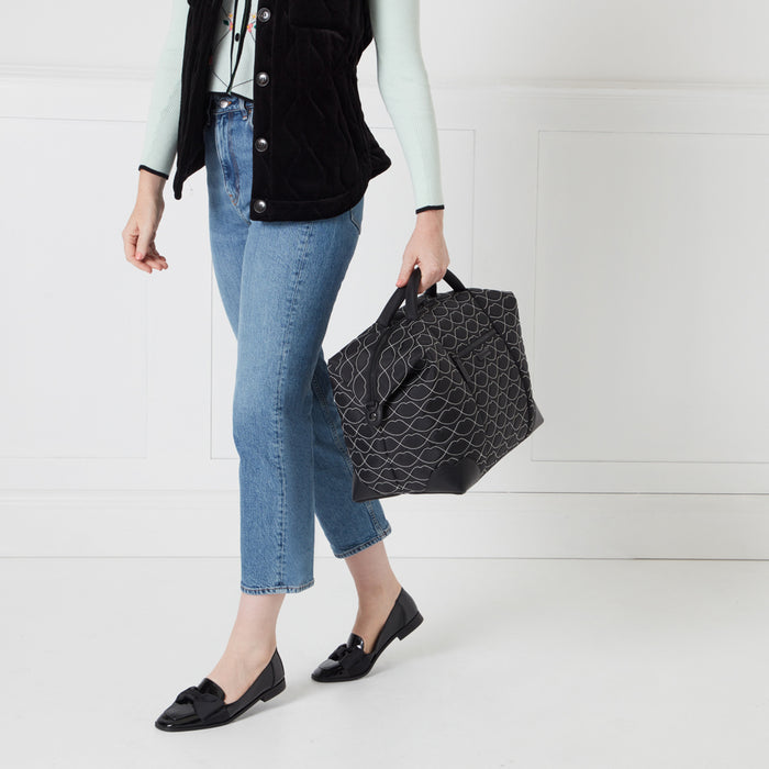 Woman in blue jeans and black vest carrying a black and white patterned handbag.