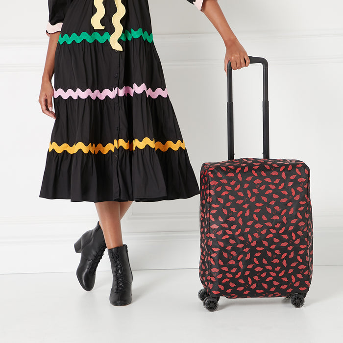 A woman in a black dress with colorful trim stands next to a suitcase covered in a red lips pattern on a white background.