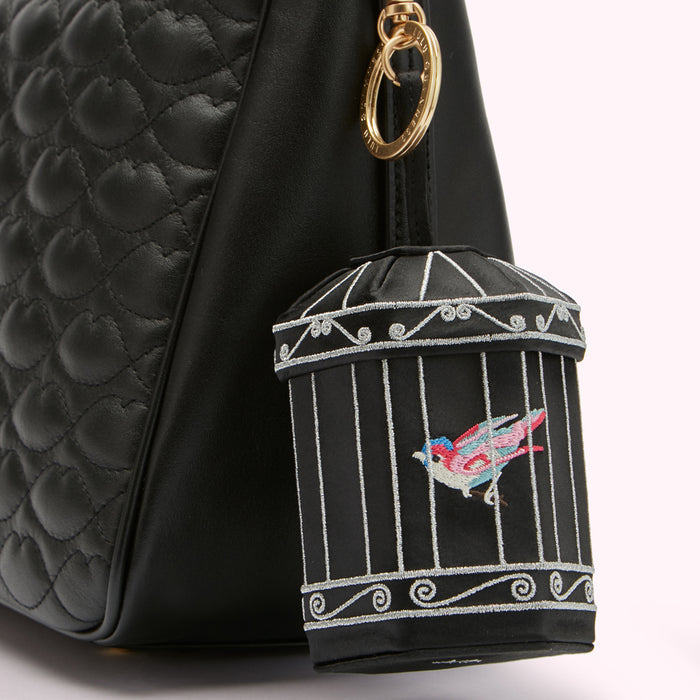 Close-up of a black quilted handbag with a birdcage-shaped charm attached. The charm is black with silver embroidery depicting a bird inside.