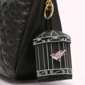 Close-up of a black quilted handbag with a birdcage-shaped charm attached. The charm is black with silver embroidery depicting a bird inside.
