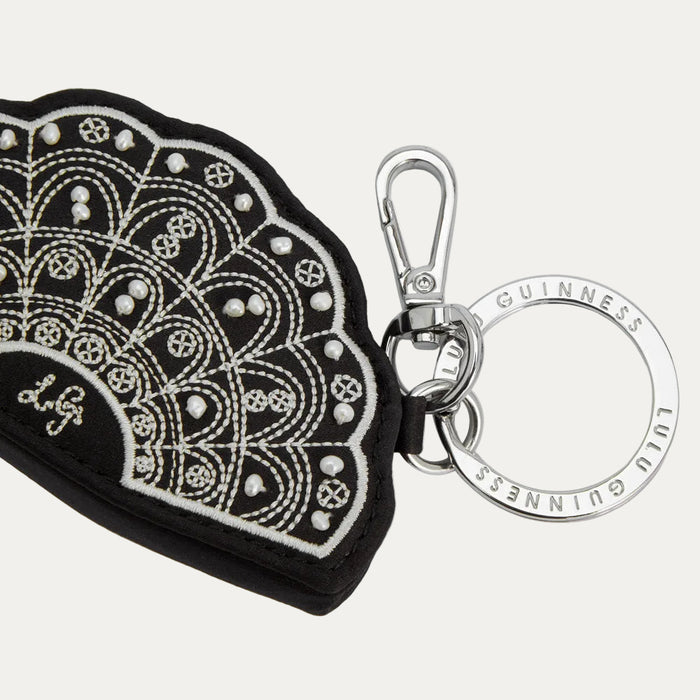 Black leather fan-shaped keyring with white embroidery and faux pearls, attached to a silver keyring with "Lulu Guinness" engraved on it.