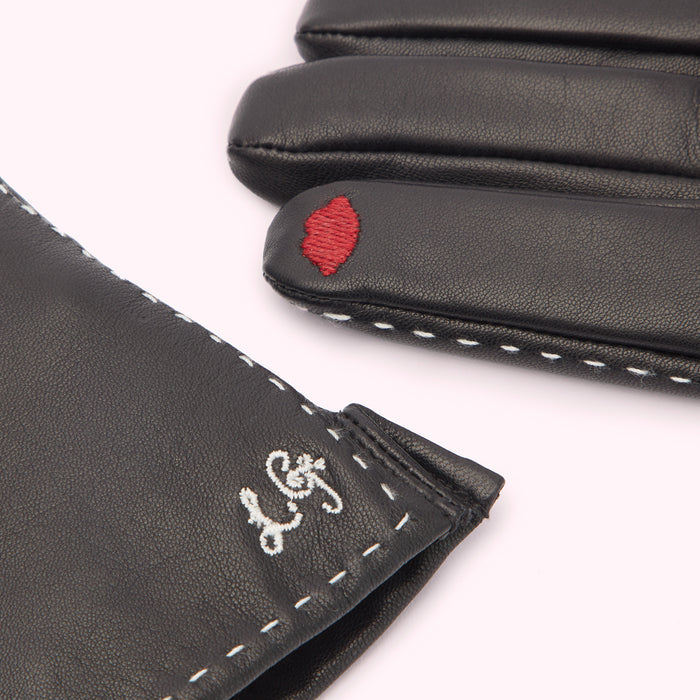 Close-up of a pair of black leather gloves with white stitching and embroidered details: a red lip on one glove and a logo on the other.