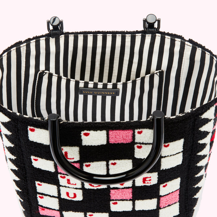 Interior view of a Lulu Guinness tote bag against a white background. The bag is black with a white and pink scrabble tile pattern and black handles. The interior lining is black and white striped.