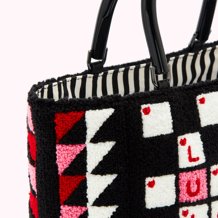Close-up of a black tote bag with a textured, geometric design in red, pink, and white against a white background. The bag has black handles.