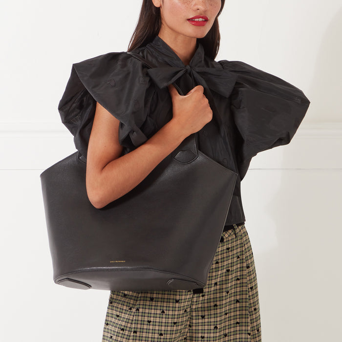 A woman with long dark hair holds a large black leather tote bag in front of her. The bag has a subtle logo on the bottom. She's wearing a black, puffy-sleeved blouse with a bow at the neck, and a plaid skirt. The background is white.