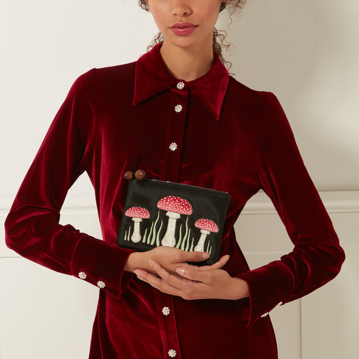 Woman in a burgundy velvet dress holding a black silk pouch embroidered with red mushrooms against a white background.