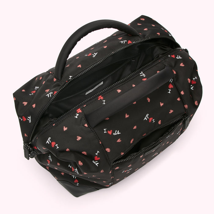 Overhead view of a black nylon duffel bag with a repeating pattern of red hearts and "I love Lulu" text; the bag is open, showing its interior.