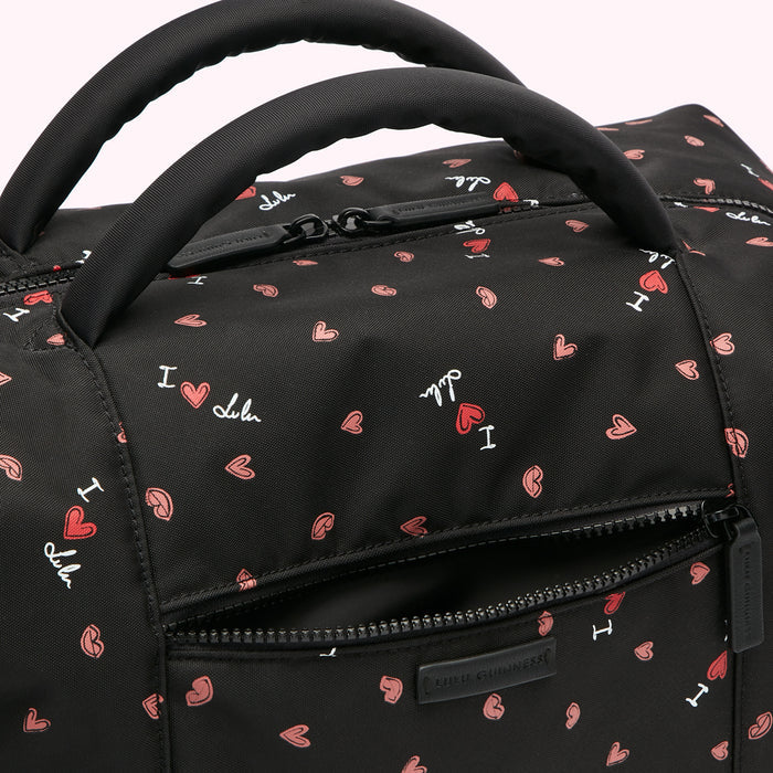 Close-up of a black Lulu Guinness duffel bag with a pink and white heart print and the words "I love Lulu" repeated on it.