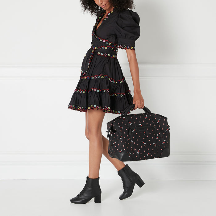 A woman in a black eyelet dress and black ankle boots carries a black weekend bag with a heart print.