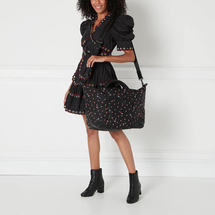 Woman in black dress holding black weekend bag with pink hearts pattern against white background.