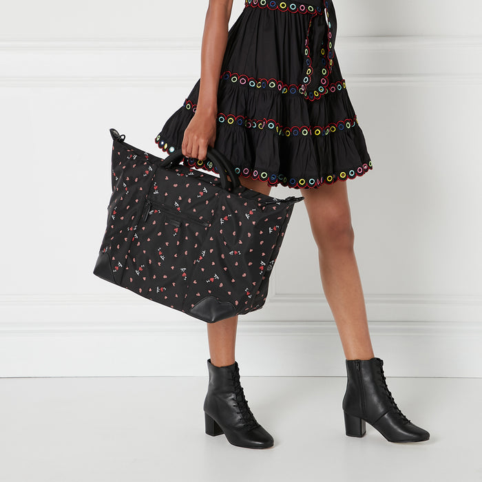 A woman in a black dress holds a black nylon tote bag with a multicolored "I love NY" print. She is wearing black lace-up ankle boots. The background is white.
