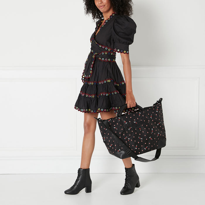 A woman in a black dress and black ankle boots carries a large black tote bag with a pink and red heart pattern against a white background.