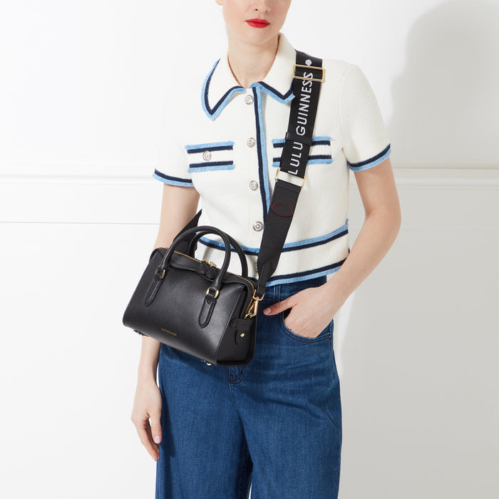 Woman in a white cardigan and blue jeans is modelling a black Lulu Guinness handbag with a black and white logo strap.