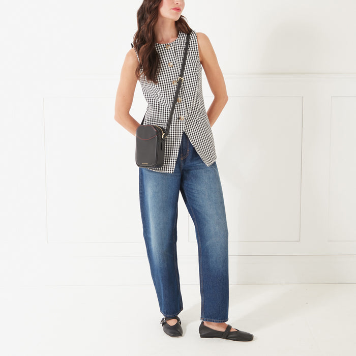 Full length image of a model with dark hair wearing a black and white gingham button up top, dark denim jeans and a black leather crossbody bag with contrasting red lip shaped stitching on the front pocket.
