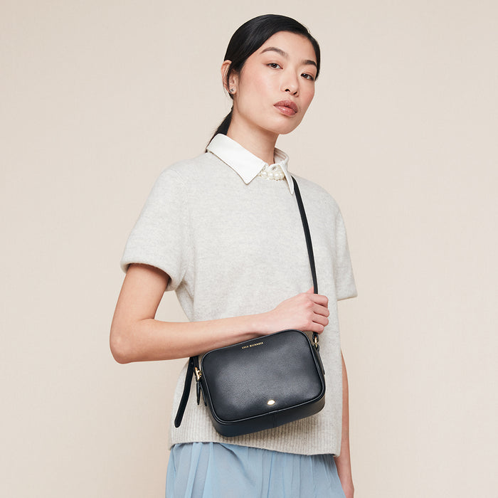 Women in a grey short sleeve jumper wearing a black leather crossbody bag against a beige background.