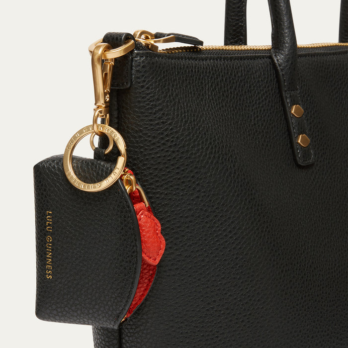 Close-up of a black pebbled leather handbag with gold-tone hardware, a matching black and red leather key pouch embossed with 'LULU GUINNESS', shown against a light neutral background.