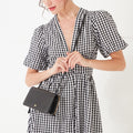 A close-up image of a model with dark hair wearing a black and white gingham dress and a black leather crossbody bag with a gold lip shaped pin on the front.