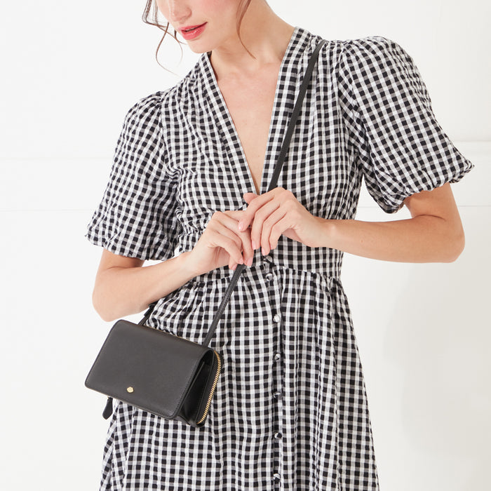 A close-up image of a model with dark hair wearing a black and white gingham dress and a black leather crossbody bag with a gold lip shaped pin on the front.