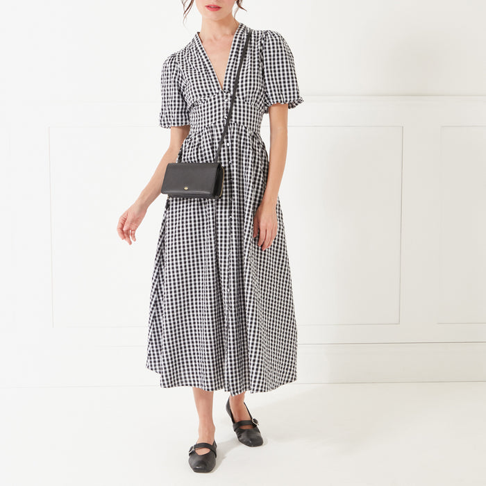 A full-length image of a model with dark hair wearing a black and white gingham dress and a black leather crossbody bag with a gold lip shaped pin on the front.