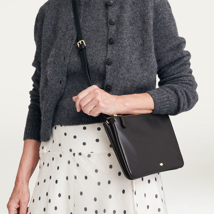 Person wearing a gray cardigan and polka dot skirt holding a black handbag against a plain background