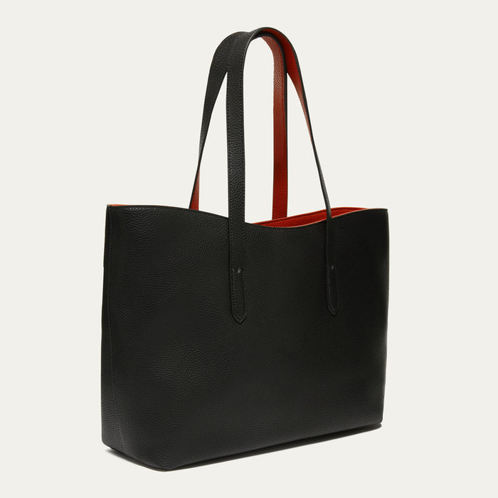 A black pebbled leather tote bag with long shoulder straps, featuring a vibrant red interior lining visible along the top edge, set against a plain off-white background.