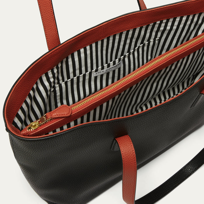 Close-up of a black pebbled leather tote bag with red leather trim and handles, showing a black and white striped interior lining and a gold zipper.