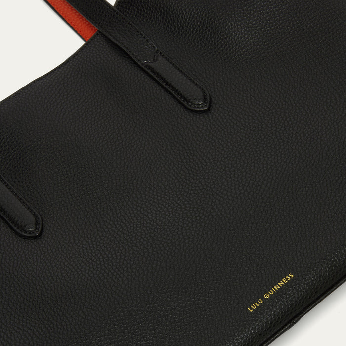 Close-up of a black pebbled leather tote bag with gold 'LULU GUINNESS' branding on the bottom right, a red accent strap at the top, and black leather handle tabs on a white background.