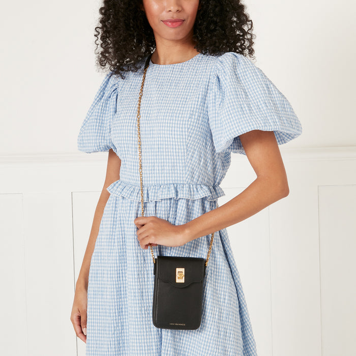 A woman in a light blue gingham dress wears a black Lulu Guinness crossbody phone bag with a gold chain strap.