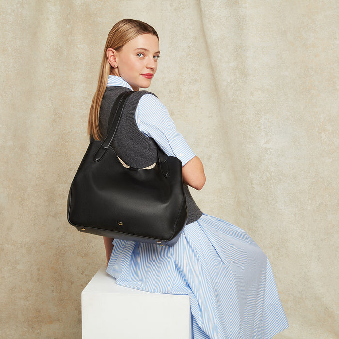 Woman sitting on a plinth holding a large black leather shoulder bag against a beige background