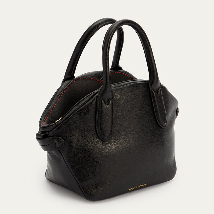 An angled side image of a black leather handbag with a top handle and a gold foil Lulu Guinness logo. Revealing contrasting red stitching on the top.