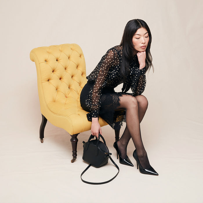 Woman sitting on a yellow chair holding a black handbag against a beige background